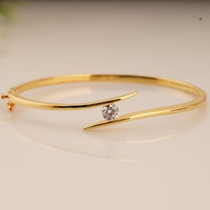 Puede incluir: Una pulsera de brazalete dorada con una sola gema redonda y transparente. La pulsera tiene un diseño elegante y moderno, con la gema sujeta entre dos extremos curvos y puntiagudos. El cierre es visible en el lado izquierdo.