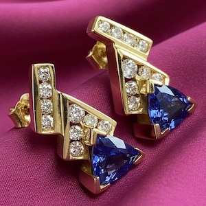 May include: A pair of gold earrings with a geometric design. Each earring features a cluster of round diamonds and a triangular, deep blue gemstone. The earrings are set against a pink background.