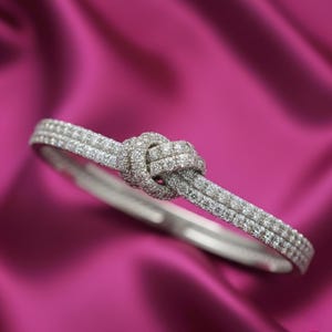 Puede incluir: Una pulsera de plata con diseño de nudo, incrustada con numerosas piedras pequeñas y brillantes. La pulsera se presenta sobre un fondo rosa vibrante, resaltando sus intrincados detalles y su estilo elegante.