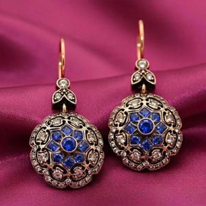 May include: A pair of ornate drop earrings with a floral design. The earrings feature a gold-tone metal setting, with a central cluster of blue gemstones surrounded by smaller white gemstones. The earrings have a lever back closure.