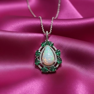 Puede incluir: Un collar de plata con un colgante en forma de pera. El colgante presenta un ópalo blanco grande rodeado de pequeñas esmeraldas verdes y un halo de piedras preciosas transparentes. El collar se muestra sobre una tela de satén rosa.
