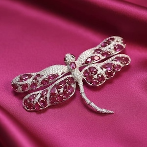 Puede incluir: Broche de libélula enjoyada con detalles de rubíes y diamantes. Las alas están adornadas con gemas de rubí, mientras que el cuerpo y los bordes están incrustados con diamantes. Sobre un fondo de satén rosa.