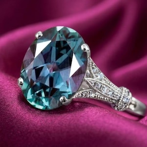 May include: A silver ring with a large, oval-cut gemstone. The gemstone has a teal and blue color, with facets that catch the light. The ring's band is detailed with small, clear stones and intricate metalwork, set against a deep pink background.