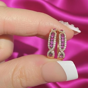 May include: A pair of gold hoop earrings with a twisted design. Each earring features a row of pink gemstones and is encrusted with small white gemstones. The earrings are held against a pink satin background.