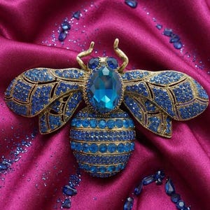 May include: A detailed bee brooch, adorned with blue gemstones and gold-toned metal. The brooch has a large central blue stone and smaller gems covering the wings and body, set against a magenta fabric background.