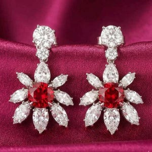 Puede incluir: Un par de pendientes deslumbrantes con un diseño floral. Cada pendiente tiene una gran piedra preciosa blanca redonda con talla brillante en la parte superior, con piedras más pequeñas debajo. El punto focal es una vibrante piedra preciosa roja rodeada de piedras blancas en forma de pétalo.