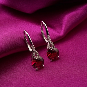 Puede incluir: Un par de pendientes colgantes plateados con una piedra preciosa roja oscura en forma de lágrima. Los pendientes presentan un pequeño acento brillante encima de la piedra preciosa y un cierre de palanca. Se exhiben sobre una tela de color magenta vibrante.