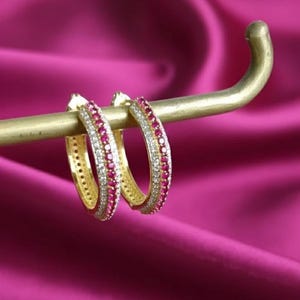 Könnte beinhalten: Ein Paar goldene Creolen mit abwechselnden Reihen roter und klarer Edelsteine. Die Ohrringe werden auf einem goldenen Stab vor einem leuchtend rosa Hintergrund präsentiert, der ihr funkelndes Design hervorhebt.