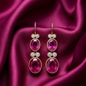 May include: A pair of gold drop earrings featuring two oval-cut vibrant pink gemstones each, accented by small round clear stones set in gold. The earrings have a hook-style closure and are displayed against a deep magenta satin fabric.