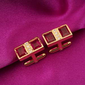 May include: A pair of gold-toned cufflinks with rectangular ruby-red gemstones and small, clear accent stones. The cufflinks have a classic design and are set against a deep magenta background.