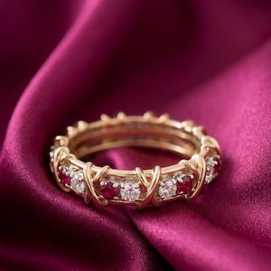 May include: A gold band ring featuring an alternating pattern of round-cut red rubies and clear white diamonds. The metal setting incorporates a repeating X-shaped design that secures each gemstone individually around the entire circumference.