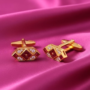 May include: A pair of gold cufflinks with a geometric design. Each cufflink features a central red gemstone surrounded by smaller clear stones. The cufflinks are set against a pink satin background.