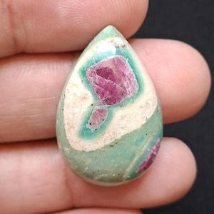 Puede incluir: Una piedra preciosa en forma de lágrima con una mezcla de colores. La piedra presenta un área central de color rosa intenso, rodeada de blanco y bandas de color verde claro. La forma general es suave y pulida, con un aspecto natural y orgánico.