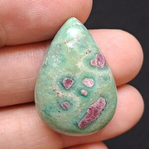 Puede incluir: Una gema en forma de lágrima con una base de color verde claro y parches irregulares de rosa intenso y rojo. La piedra es lisa y pulida, con una apariencia ligeramente translúcida. La gema se sostiene en una mano sobre un fondo oscuro.