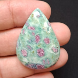 Puede incluir: Una gema en forma de lágrima con una base verde claro y parches dispersos de color rojo intenso. La piedra tiene una superficie pulida y un aspecto natural. La gema se sostiene en una mano.