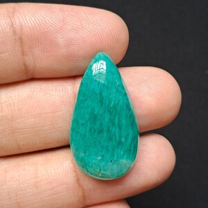 Op de afbeelding: Een gepolijste, druppelvormige edelsteen in tinten van blauwgroen en turkoois. De steen heeft een glad, glanzend oppervlak met subtiele adering. De edelsteen wordt in een hand tegen een donkere achtergrond gehouden.