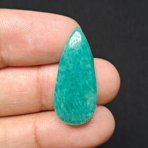 Op de afbeelding: Een gepolijste, druppelvormige edelsteen in tinten van blauwgroen en turkoois. De steen heeft een glanzende afwerking en zichtbare interne patronen. De edelsteen wordt in een hand tegen een donkere achtergrond gehouden.