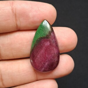 Op de afbeelding: Een druppelvormige edelsteen met een gepolijst oppervlak. Het bovenste gedeelte is diepgroen, overgaand in een rijke, donkerrood-paarse tint. De steen wordt in een hand gehouden tegen een donkere achtergrond.