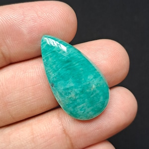 Op de afbeelding: Een gepolijste, druppelvormige edelsteen in tinten van blauwgroen en turkoois. De steen heeft een glad, glanzend oppervlak met zichtbare natuurlijke patronen. De edelsteen wordt in een hand tegen een donkere achtergrond gehouden.