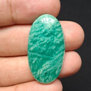 Op de afbeelding: Een ovale edelsteen met een gepolijst oppervlak. De steen heeft een levendige turquoise kleur met een patroon van witte en licht turquoise strepen. De edelsteen wordt in een hand tegen een donkere achtergrond gehouden.