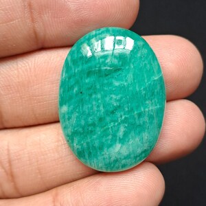 Op de afbeelding: Een gepolijste, ovaalvormige edelsteen in tinten groen en blauwgroen. De steen heeft een glad, reflecterend oppervlak met natuurlijke aderingpatronen. De edelsteen wordt in een hand gehouden tegen een donkere achtergrond, wat de kleur en textuur benadrukt.