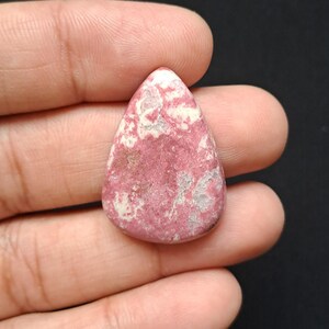 Op de afbeelding: Een druppelvormige edelsteen met een roze en wit gevlekt patroon. De gepolijste steen heeft een glad oppervlak en wordt in een hand tegen een donkere achtergrond gehouden. De kleur van de steen is een mix van diep roze en wit.