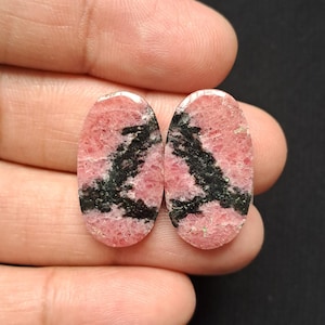 Op de afbeelding: Twee ovale edelstenen met een roze basiskleur en zwarte adering. De stenen worden in een hand gehouden tegen een donkere achtergrond. De edelstenen hebben een gepolijst oppervlak. Elke steen is ongeveer 2 cm lang.