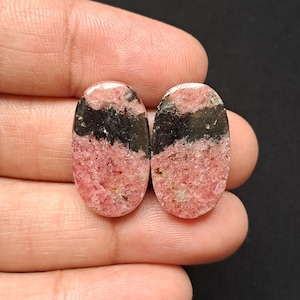 Op de afbeelding: Twee ovale edelstenen met een roze en zwart kleurenschema. De edelstenen hebben een gepolijst oppervlak en een textuur. De roze secties hebben een gespikkeld uiterlijk, terwijl de zwarte secties effen zijn. De edelstenen worden in een hand gehouden.