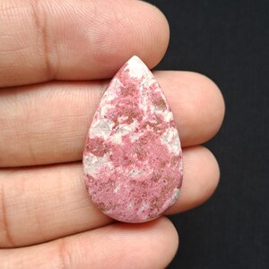 Op de afbeelding: Een druppelvormige edelsteen met een roze en wit gevlekt patroon. De gepolijste steen heeft een glad oppervlak en wordt in een hand gehouden tegen een donkere achtergrond. De kleur van de edelsteen is een mix van lichtroze, wit en donkerder roze.