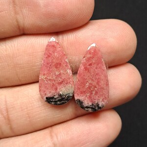 Op de afbeelding: Twee druppelvormige edelstenen in roze en zwart. De edelstenen hebben een textuur en worden in een hand gehouden. Roze is de hoofdkleur, met zwart aan de basis van de druppelvorm. Waarschijnlijk voor sieraden.