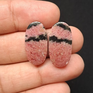 Op de afbeelding: Twee ovale rhodochrosiet edelstenen. De stenen hebben een roze basiskleur met zwarte banden en insluitsels. De edelstenen zijn gepolijst en hebben een glad oppervlak. De achtergrond is donker.