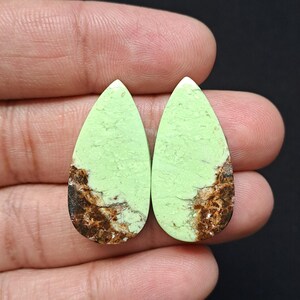 Op de afbeelding: Twee peervormige edelstenen met een lichtgroene tint en bruine marmering. De stenen worden in een hand gehouden tegen een zwarte achtergrond. De edelstenen zijn waarschijnlijk voor het maken van sieraden.