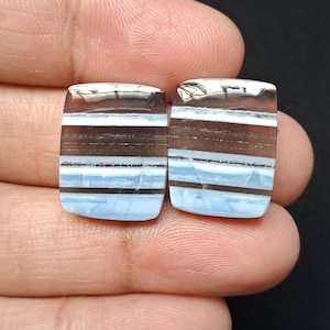 Op de afbeelding: Twee rechthoekige edelstenen met afgeronde hoeken. De stenen hebben een gelaagd patroon van bruine, witte en lichtblauwe banden. De edelstenen worden in een hand tegen een zwarte achtergrond gehouden.