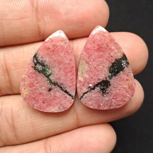 Op de afbeelding: Twee druppelvormige edelstenen met een roze tint, met zwarte en groene adering. De stenen hebben een textuur en worden in een hand tegen een donkere achtergrond gehouden. De edelstenen zijn waarschijnlijk voor het maken van sieraden.