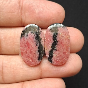 Op de afbeelding: Twee ovale edelstenen met een roze en zwart patroon. De edelstenen worden in een hand gehouden tegen een donkere achtergrond. Roze is de dominante kleur, met zwarte strepen die door de stenen lopen.