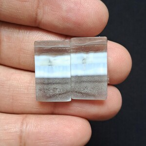 Op de afbeelding: Twee rechthoekige edelstenen met een gelaagde uitstraling. De lagen omvatten witte, lichtblauwe en grijsbruine tinten. De stenen worden in een hand tegen een donkere achtergrond gehouden.