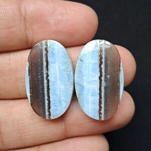 Op de afbeelding: Twee ovale edelstenen. Elke steen heeft een lichtblauw midden met een rasterpatroon, begrensd door een verticale band van bruin en wit. De stenen worden in een hand tegen een zwarte achtergrond gehouden.
