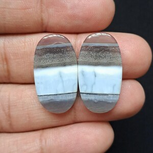 Op de afbeelding: Twee ovale edelstenen met een gelaagd patroon. De bovenste laag is lichtbruin, gevolgd door een grijze en witte band en een lichtblauwe sectie. De onderkant is donkerder grijs. De stenen worden in een hand gehouden.