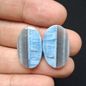 Op de afbeelding: Twee ovale edelstenen, elk verdeeld in twee secties: een lichtblauwe sectie en een grijze sectie. Een donkerdere grijze streep loopt door de grijze sectie. De edelstenen worden in een hand tegen een zwarte achtergrond gehouden.