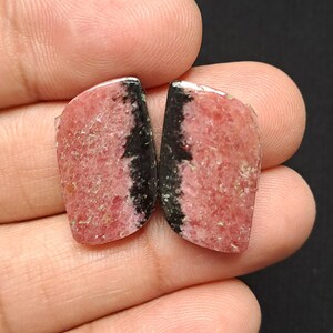 Op de afbeelding: Twee gepolijste edelstenen, elk met een mix van roze en zwart. De roze gebieden hebben een gespikkelde textuur, terwijl de zwarte secties effen zijn. De stenen worden in een hand gehouden.