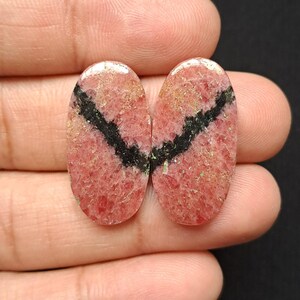 Op de afbeelding: Twee ovale edelstenen met een rozerode tint en een opvallende zwarte streep. De stenen hebben een getextureerd oppervlak en een gepolijste afwerking, die natuurlijke patronen onthult. De edelstenen worden in een hand gehouden.