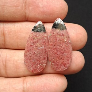 Op de afbeelding: Twee druppelvormige edelstenen met een rozerode tint en een textuur. De bovenkant van elke steen heeft een zilverkleurige metalen dop. De edelstenen worden in een hand gehouden tegen een donkere achtergrond.