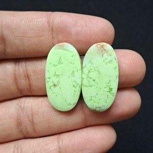 Op de afbeelding: Twee ovale edelstenen met een lichtgroene tint en bruine accenten. De stenen hebben een glad, gepolijst oppervlak met natuurlijke aderingpatronen. De edelstenen worden in een hand gehouden tegen een donkere achtergrond, wat hun kleur en textuur benadrukt.