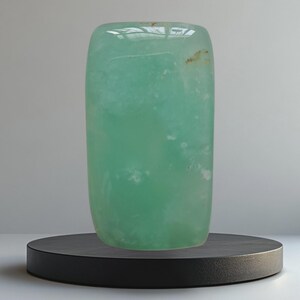 Op de afbeelding: Een grote, gepolijste, rechthoekige edelsteen in een lichtblauwe kleur. De steen heeft een glad, reflecterend oppervlak en staat op een zwarte, cirkelvormige sokkel. De edelsteen lijkt doorschijnend, met subtiele interne patronen.