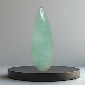 Op de afbeelding: Een grote, gepolijste, druppelvormige edelsteen in een lichtgroene tint. De edelsteen wordt getoond op een kleine, ronde, zwarte sokkel tegen een neutrale grijze achtergrond. Het oppervlak van de steen reflecteert licht, wat de gladde textuur benadrukt.