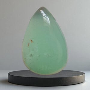 Op de afbeelding: Een grote, druppelvormige edelsteen in een lichtgroene tint. De gepolijste steen heeft een doorschijnende kwaliteit, met subtiele insluitsels en een glanzend oppervlak. Hij staat op een kleine, ronde, zwarte sokkel, wat een opvallend contrast creëert.