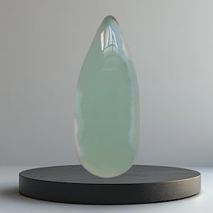 Op de afbeelding: Een grote, druppelvormige sculptuur in een lichtgroene tint, gepresenteerd op een zwarte, cirkelvormige sokkel. De sculptuur heeft een glad, glanzend oppervlak dat licht reflecteert. Het ontwerp is minimalistisch en modern, geschikt voor interieurdecoratie.