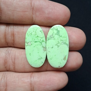 Op de afbeelding: Twee ovale edelstenen in een lichtgroene kleur met een gemarmerd patroon. De stenen hebben een gepolijst oppervlak en worden in een hand gehouden tegen een donkere achtergrond. Deze edelstenen zijn waarschijnlijk voor het maken van sieraden.