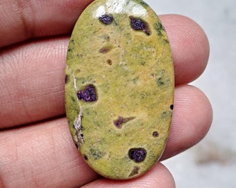 May include: Oval-shaped gemstone with a light green base color and dark purple inclusions. The polished stone has a smooth surface with a few small imperfections. The gemstone is held in a hand, showcasing its natural beauty and unique pattern.