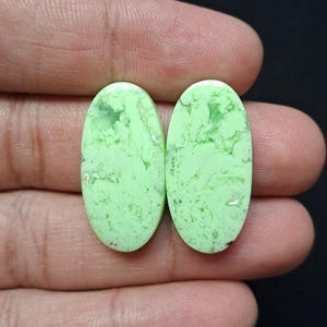 Op de afbeelding: Twee ovaalvormige, lichtgroene edelstenen met een gemarmerd uiterlijk. De stenen hebben een gepolijst oppervlak en zijn geplaatst tegen een donkere achtergrond. De edelstenen zijn ongeveer 2,5 cm lang.
